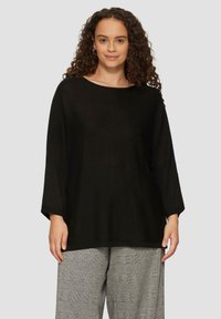 Zwarte gebreide top met een brede halslijn en driekwartmouwen, met een losse pasvorm. G gedragen met grijze geruite broek.