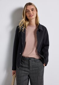 Giacca nera con zip argentata e bottoni automatici, indossata sopra una t-shirt rosa chiaro e pantaloni grigi a fantasia con taglio dritto.