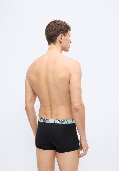 Άνδρας που στέκεται με την πλάτη στην κάμερα, φοράει μαύρα ανδρικά εσώρουχα τύπου μποξεράκια με φωτεινό γαλάζιο ζωνάρι μέσης που φέρει το λογότυπο EA.