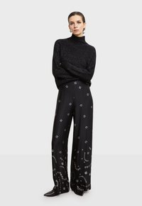 Crni pleteni pulover s visokim ovratnikom i teksturiranim završetkom, uparen s širokim crnim hlačama koje imaju srebrne geometrijske uzorke.