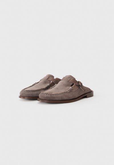 Scarpe slip-on in suede grigio con dettagli cuciti e una striscia di pelle. Presentano una suola piatta e un design a punta arrotondata.
