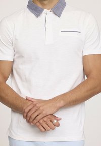 Polo à manches courtes en blanc, avec un col bleu imitant le denim et une petite poche poitrine bordée de bleu foncé. Tissu doux au toucher.