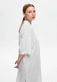 Robe blanche légère avec un col, des manches courtes ornées de détails en dentelle à œillets, et des fentes latérales pour plus de liberté de mouvement.
