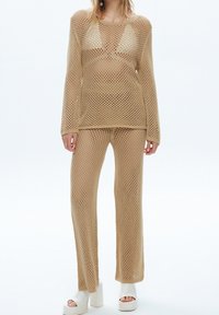 Model wearing beige open-knit sweater and matching pants over visible white undergarments, paired with white open-toe platform heels.