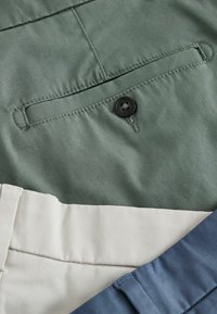 Pantalons verts, beiges et bleus pliés avec poche arrière visible et détail de bouton sur la paire verte.