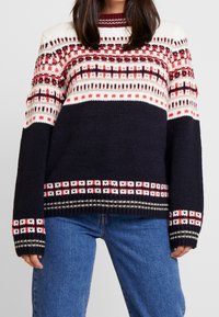 Sweter robiony na drutach w granatowym kolorze z geometrycznym wzorem w odcieniach czerwieni, bieli i czerni. Posiada okrągły dekolt, długie rękawy oraz ściągacze przy rękawach i u dołu.