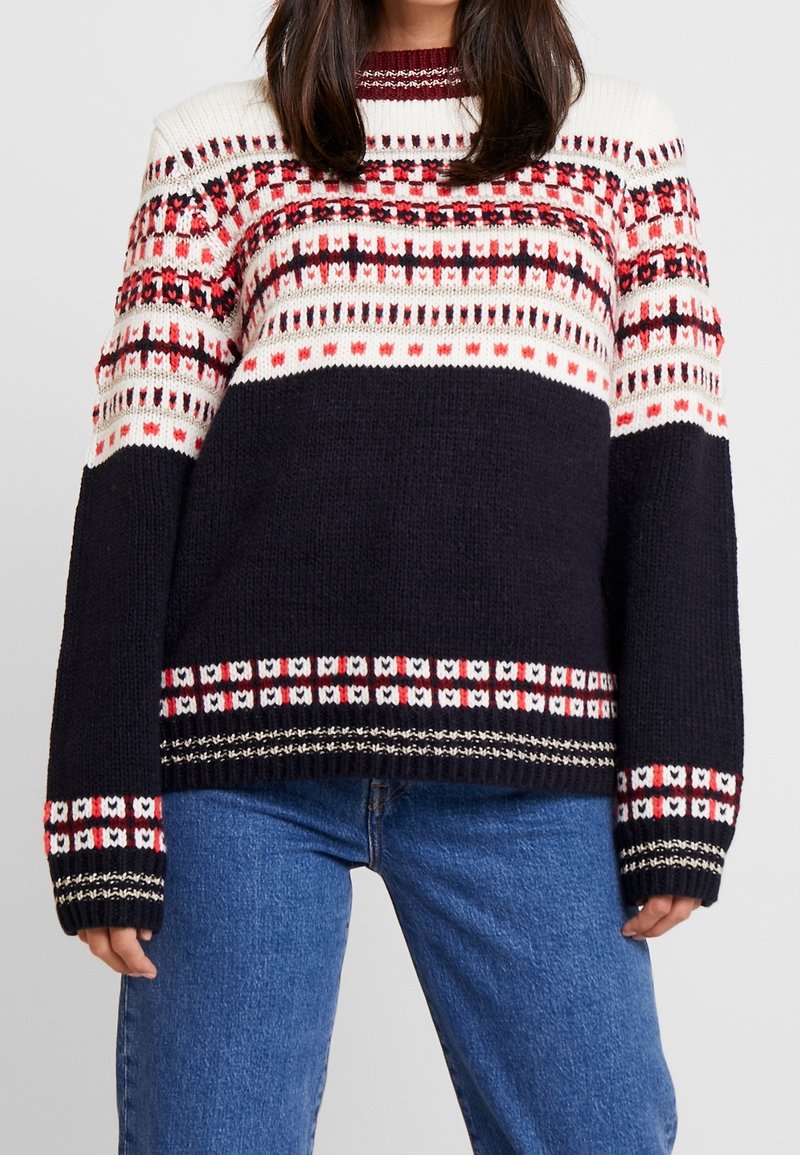 Sweter robiony na drutach w granatowym kolorze z geometrycznym wzorem w odcieniach czerwieni, bieli i czerni. Posiada okrągły dekolt, długie rękawy oraz ściągacze przy rękawach i u dołu.