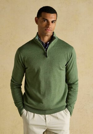 Mann trägt einen grünen Pullover mit Reißverschluss am Kragen über einem blau karierten Hemd und beigen Hosen, steht mit den Händen in den Taschen vor einem schlichten Hintergrund.