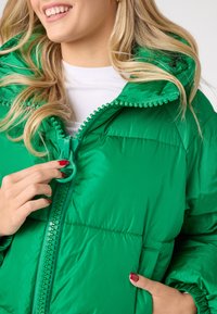 Zelena puffer jakna z visokim ovratnikom, ki ima debelo zadrgo in teksturiran rob. Gladka, sijoča tkanina s kviltanim vzorcem.