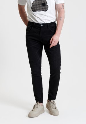 Zwarte slim-fit jeans met een zachte textuur, vijf-pocket ontwerp en lichte stiksels, gecombineerd met een wit graphic t-shirt en beige sneakers.