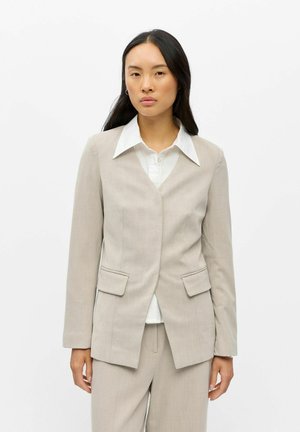 Femme aux longs cheveux foncés portant une veste de costume beige, un pantalon assorti et une chemise blanche à col, debout devant un fond blanc uni.
