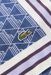 Bomullsstoff i blått med geometriske mønstre, med en grønn brodert krokodillelogo på en hvit og lilla stripete kant.