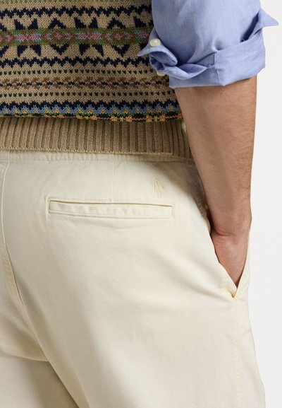 Vest tricotat cu un model geometric multicolor, asortat cu pantaloni din bumbac bej deschis, având un buzunar la spate și un detaliu subtil cu logo.