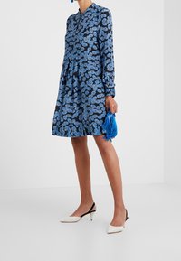 Gaun berpola bunga biru yang dikenakan oleh seorang model, menampilkan lengan panjang, kerah tinggi, dan siluet melebar. Dilengkapi dengan tas tangan rumbai biru.