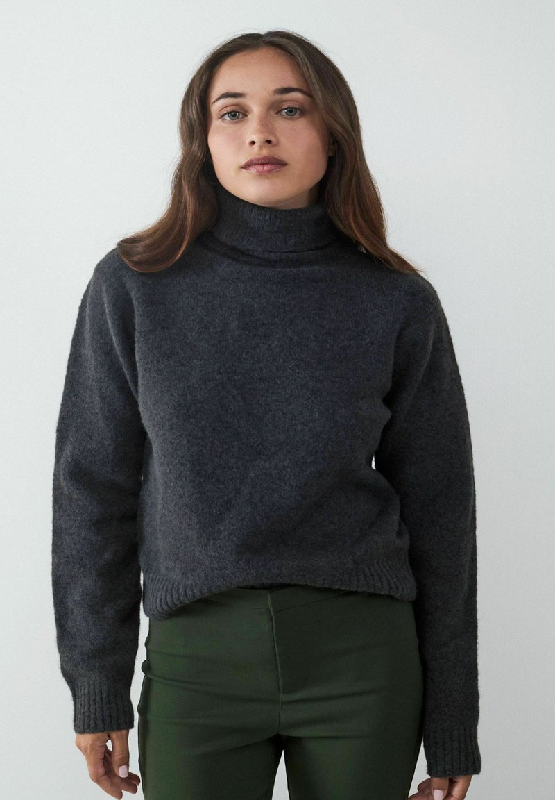 Dunkelgrauer Rollkragensweater aus weichem, strukturiertem Stoff mit gerippten Bündchen und Saum. Kombiniert mit eng anliegenden grünen Hosen.