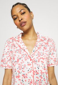 Floral short-sleeve pajama top in white, featuring a pink and black flower pattern, single pocket, and contrasting pink piping along the collar.