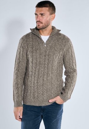 Kabelstrick, mit Reißverschluss, in hellbraun, mit hohem Kragen und strukturiertem Muster; kombiniert mit dunkelblauen Jeans.
