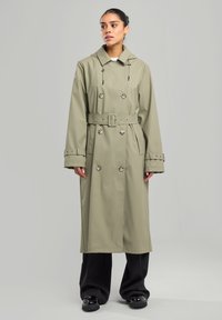 Khaki veden kestävä trench-takki, jossa on kaksiriviset napit, säädettävä nyörihuppu, vyötärövyö ja sivutaskut. Mustat leveälahkeiset housut.