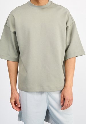 Mężczyzna noszący luźną, krótką, jasnoszarą bluzę oversize i jasnoszare szorty z teksturą, stojący na jednolitym tle.