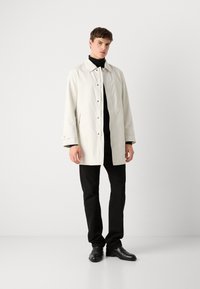 Chaqueta de un color beige claro con collar y cierre de botón, con mangas largas y una textura suave. Combinada con pantalones y zapatos negros.