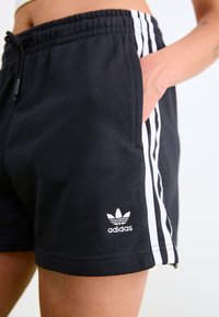 Black shorts with white Adidas logo, elastic waistband, side pockets, and three white vertical stripes on each side. Cotton blend material.