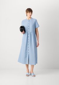 Vestito blu chiaro con pois, maniche corte, frontale con bottoni e gonna a strati. La modella tiene una pochette nera e indossa sandali blu chiaro.
