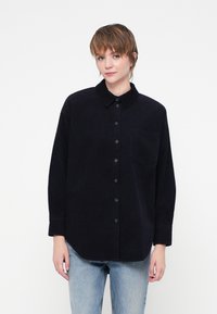 Cămașă neagră din catifea cu nasturi, guler, buzunar frontal și mâneci lungi, asortată cu blugi de culoare bleu deschis. Textură netedă, design casual.