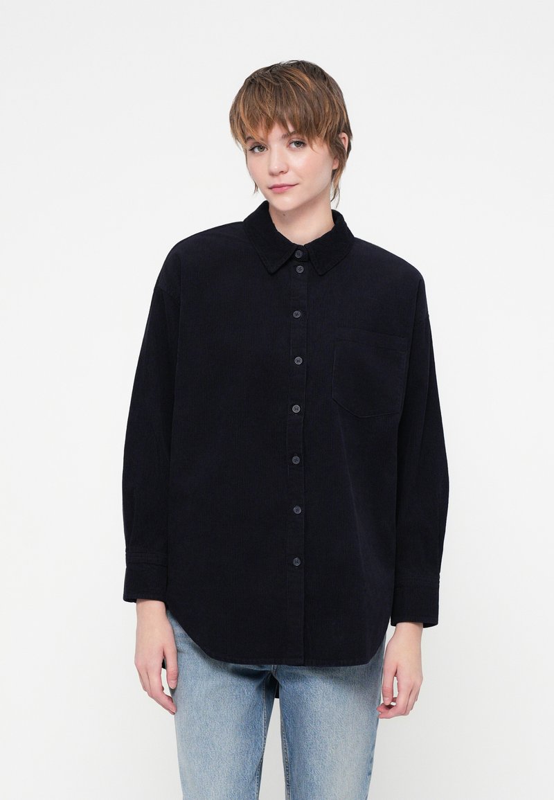 Cămașă neagră din catifea cu nasturi, guler, buzunar frontal și mâneci lungi, asortată cu blugi de culoare bleu deschis. Textură netedă, design casual.
