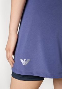 Vijolična športna majica z gladko teksturo, ki ima bel logo na robu. Majica je oblečena čez črne kratke hlače.