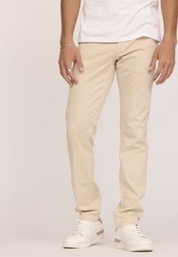 Chinos beiges en tissu lisse, avec une coupe slim et des poches standard, associés à des baskets blanches avec des accents dorés.