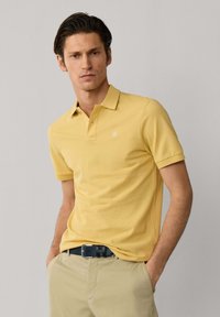 Homme portant un polo à manches courtes jaune moutarde rentré dans un pantalon beige avec une ceinture noire, debout devant un fond clair uni.
