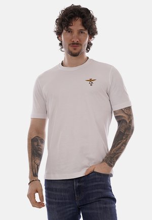 Uomo con capelli ricci scuri e tatuaggi su entrambe le braccia, indossa una maglietta bianca con un piccolo emblema e jeans blu, in piedi davanti a uno sfondo bianco.