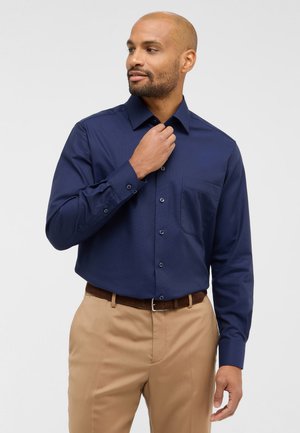 Camicia a maniche lunghe blu navy in tessuto strutturato con taschino sul petto e chiusura a bottoni. Abbinata a pantaloni beige e cintura marrone.