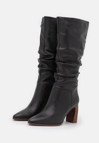 Bottes hautes jusqu'au genou en cuir noir avec un design décontracté, bouts pointus et talon bloc marron. Texture lisse avec des plis visibles.