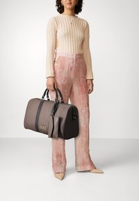 Sac à main à motifs marron avec des accents noirs et des poignées courtes, associé à un pantalon taille haute en velours rose et un pull en crème à côtes.