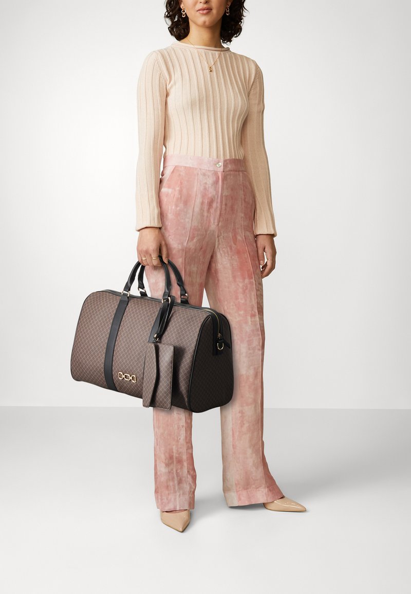 Sac à main à motifs marron avec des accents noirs et des poignées courtes, associé à un pantalon taille haute en velours rose et un pull en crème à côtes.