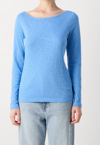 Niebieska bluzka z długim rękawem w strukturze lekkiego materiału z szerokim dekoltem. Połączona z luźnymi, jasnoniebieskimi dżinsami.
