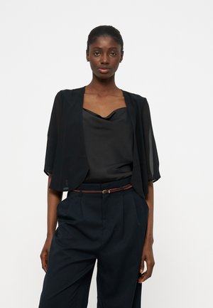 Femme à la peau foncée et aux cheveux attachés, portant une veste kimono noire transparente, un haut en satin noir et un pantalon noir avec une fine ceinture marron.