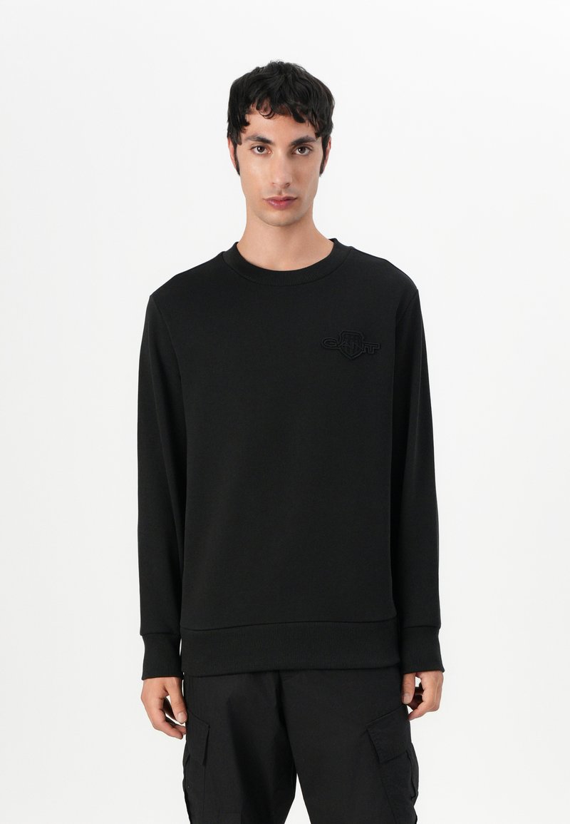 Schwarzer Pullover mit langen Ärmeln, Rundhalsausschnitt und strukturiertem Stoff. Mit einem dezenten Logo auf der Brust. Getragen mit dunklen Cargo-Hosen.