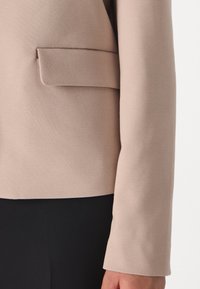 Beige blazer with a smooth texture, featuring a single chest pocket and straight-cut sleeves, paired with black fabric below.