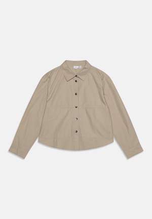 Chemise beige à manches longues et col. Présente un ourlet arrondi, deux poches avant et des boutons noirs. Tissu léger, texture lisse.