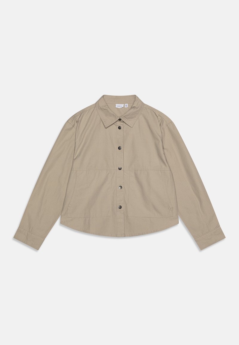 Beige overhemd met lange mouwen en een kraag. Heeft een gebogen zoom, twee voorzakken en zwarte knopen. Lichtgewicht stof, gladde textuur.