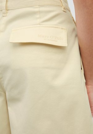 I pantaloni beige presentano una tasca posteriore con patta quadrata etichettata "Marc O'Polo", che mostra una texture del tessuto liscia e morbida.