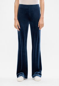 Tumši zili samta bikses ar paplašinātas formas dizainu, gludu tekstūru un elastīgu jostas daļu; apakšā saskaņotas ar baltām kedām.