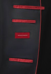 Zwarte stof met rode satijnen accenten en zakken; voorzien van een rood label met "THOMAS GOODWIN" in witte tekst. Gladde textuur, op maat gemaakt ontwerp.