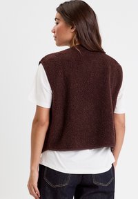 Brown fleece vest with a textured surface, sleeveless design, and rounded hem, worn over a white t-shirt and paired with dark jeans.
