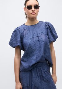 Μπλούζα σε ναυτικό μπλε με υφή και μανίκια με φουσκάλες, συνδυασμένη με φούστα στο ίδιο χρώμα. Ελαφρύ ύφασμα με υφή ζαρώματος, διακριτική λεπτομέρεια με στρώσεις.