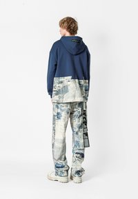 Sudadera de marina con una sección inferior texturizada en azul claro y blanco, combinada con vaqueros holgados que presentan un patrón mixto y un detalle de etiqueta lateral.