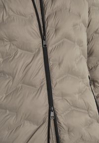 Beige pufferjack met zwarte ritsen, met een hexagonaal quiltpatroon en een gladde textuur. Ritszakken aan de zijkant zichtbaar.