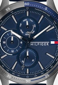 Sinine kellanägu hõbedaste tunnimarkeritega, kolm aladialki päeva, 24-tunni ja sekundite jaoks, ning "HILFIGER" logo lipuga paremal.